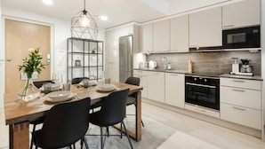 A modern kitchen with a dining area, featuring a wooden table, black chairs, a built-in bookshelf, and stainless steel appliances. - Superior Queen Room in Tammela Soccer Stadium! (Tampere)