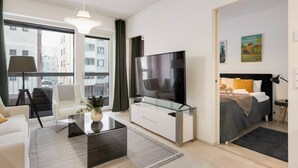 A modern hotel room with a large flat-screen TV, a sofa, a coffee table, a bed with a black headboard, and a window with curtains. - Superior Queen Room in Tammela Soccer Stadium! (Tampere)