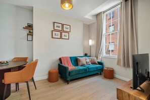 A modern living room with a teal sofa, orange chairs, a wooden dining table, and a TV on a wooden stand. - GuestReady - Stylish getaway in Liverpool (Liverpool)