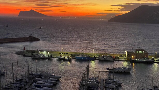 Un puerto deportivo al atardecer con barcos amarrados, un faro y montañas a lo lejos.