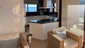 Een moderne keuken met een centraal eiland, een eetgedeelte met rieten meubels en uitzicht op zee door grote ramen.