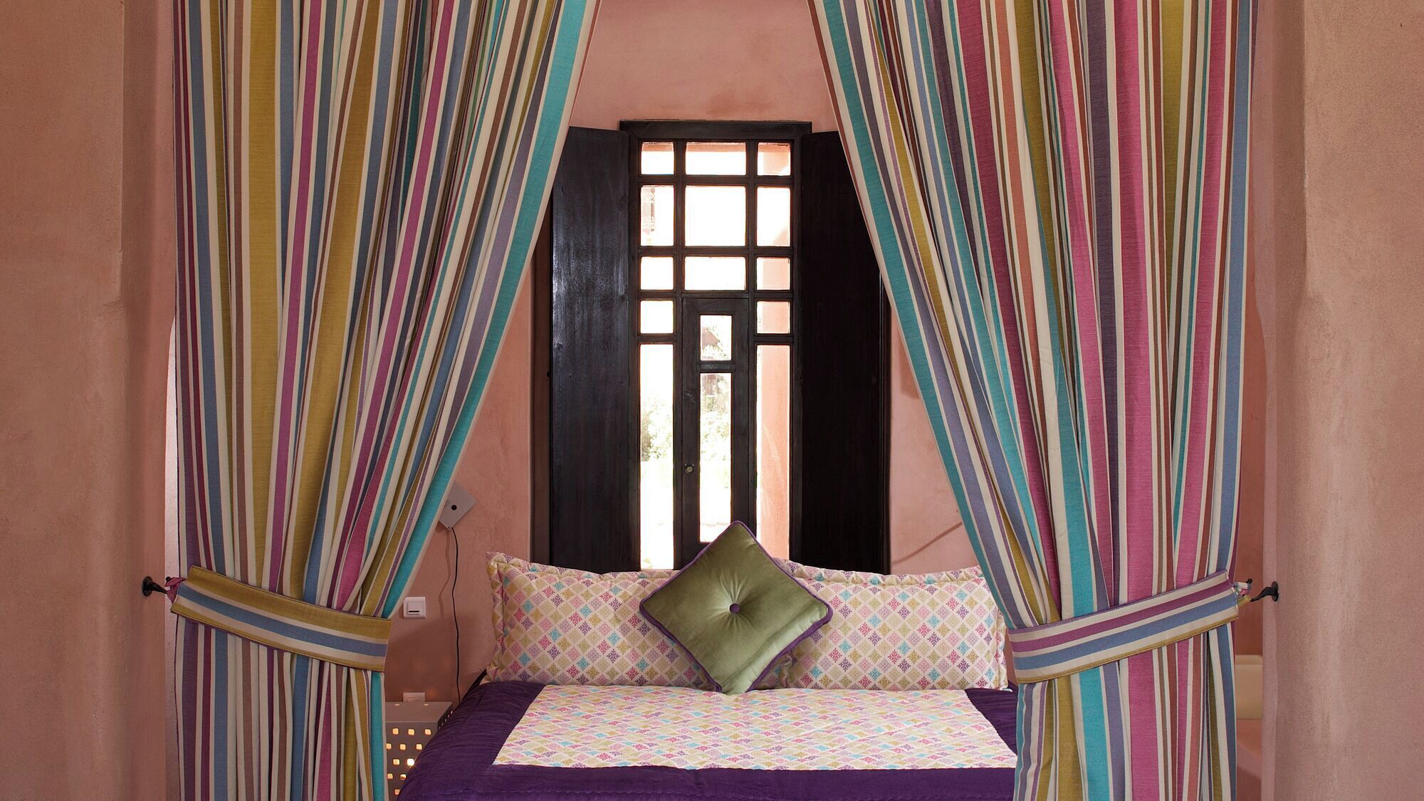 A bed with a patterned bedspread, pillows, and curtains in a room with a window and a wall socket.