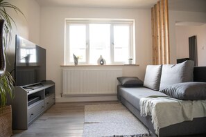 A modern living room with a grey sofa, a flat-screen TV, a radiator, and a window with curtains. - Dorset Stylish Apartment (Poole)