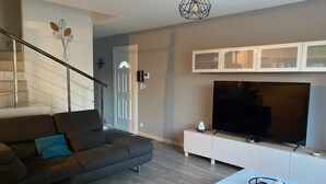 Ein modernes Wohnzimmer mit einem grauen Sofa, einem weißen Couchtisch und einem flachbildfernseher, der an einem weißen Schrank befestigt ist. Im Hintergrund ist eine Treppe mit Glastrennwand sichtbar.