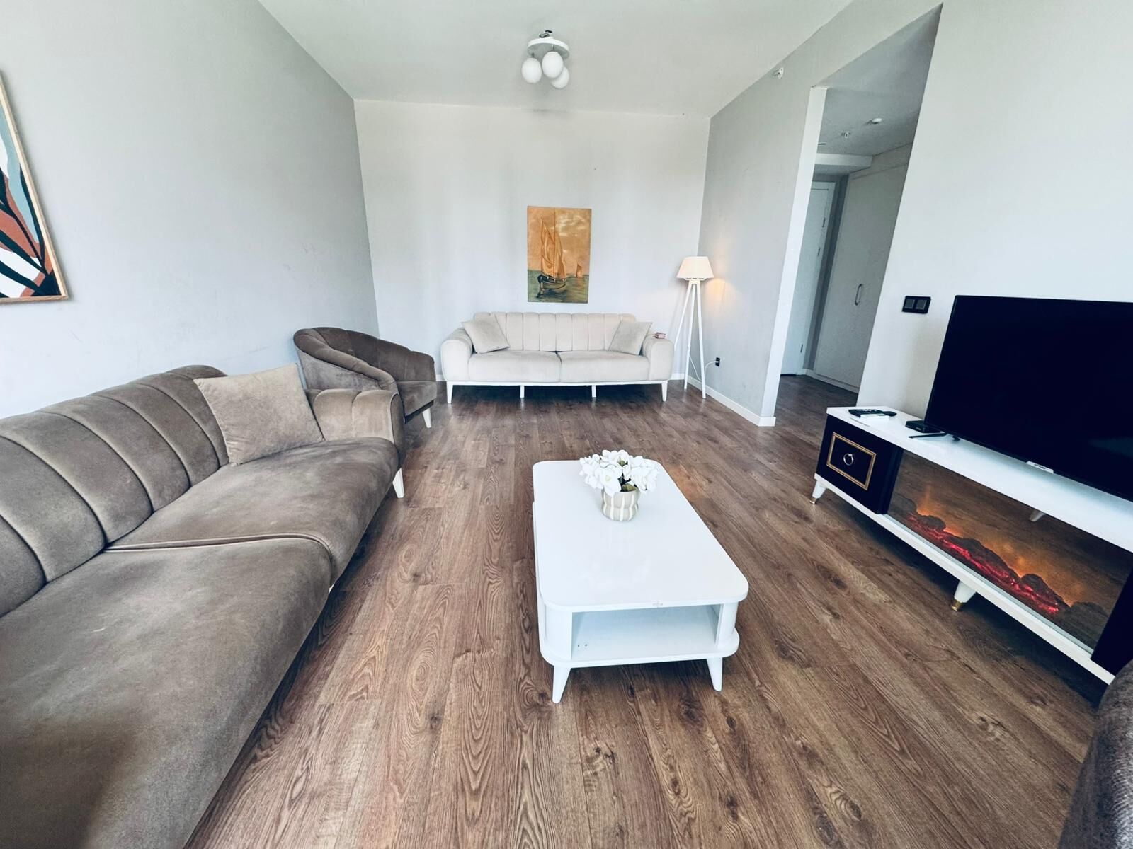 Un salon moderne avec un canapé gris, une table basse blanche ornée de fleurs, un téléviseur à écran plat et une cheminée au décor évoquant la lave.