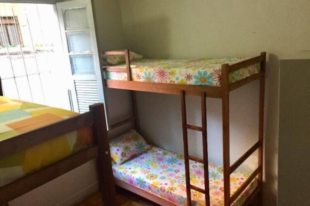 Uma cama beliche com roupa de cama florida, uma mesa de madeira e uma cadeira em um quarto com janela e porta.