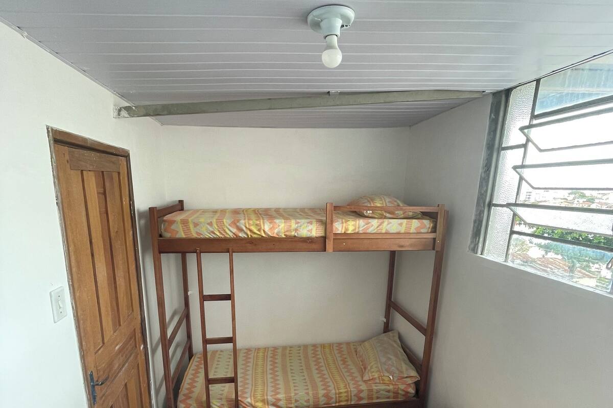 Quarto pequeno e compacto com beliches, porta de madeira e janela com grades.