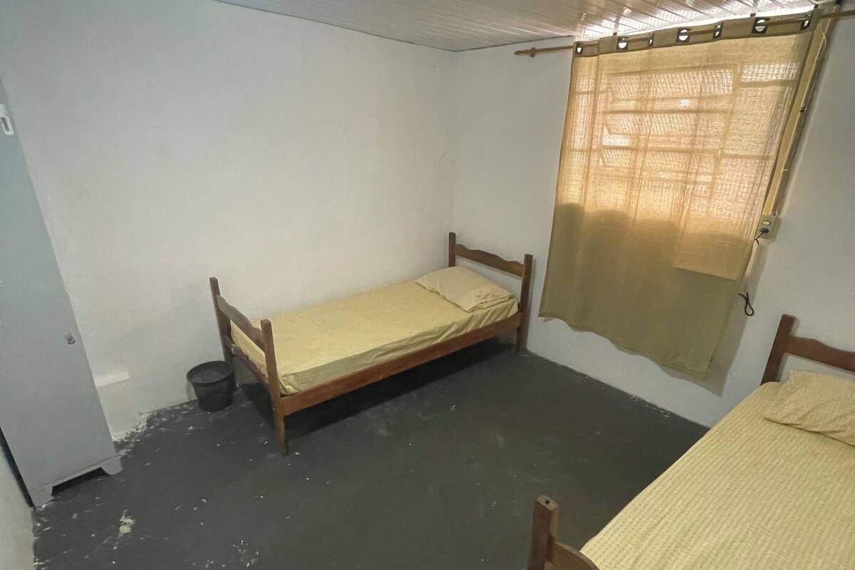 Quarto pequeno e com pouca luz, com duas camas de solteiro, um ventilador de teto e uma janela com cortina.