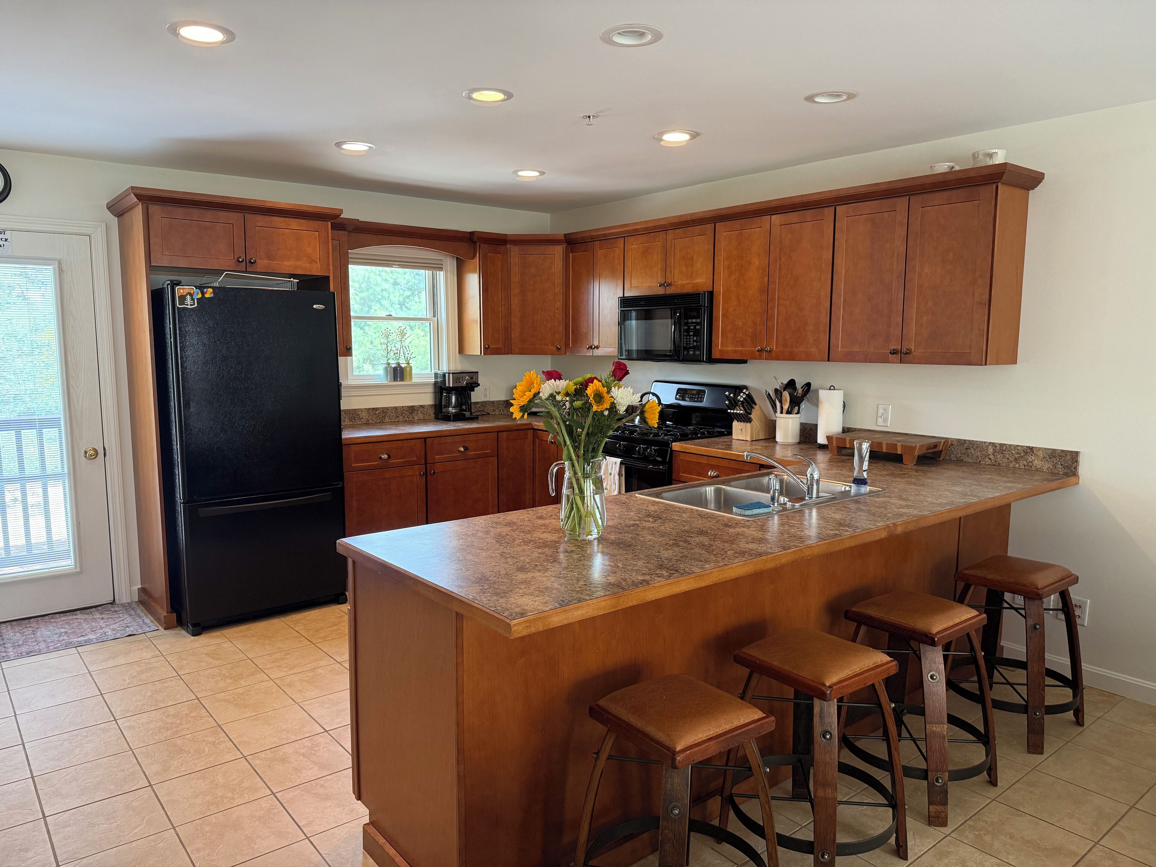 A kitchen with wooden cabinets, a black refrigerator, a stove, and a sink. There are three stools and a vase with flowers on the counter.