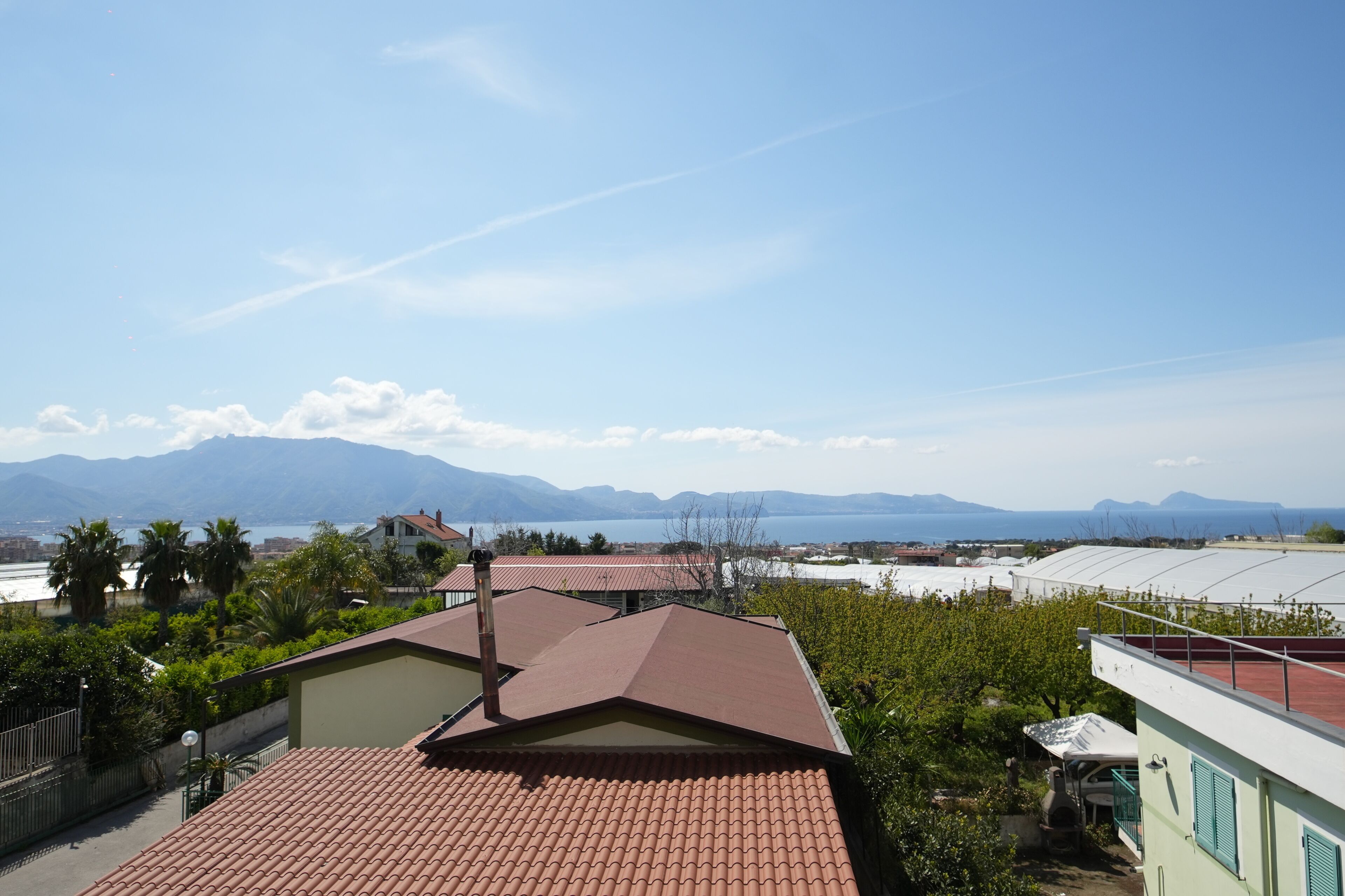 A residential area with a red-tiled roof house, a green building with a white roof, and a view of mountains and a body of water in the distance.