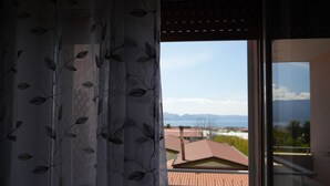 Une vue à travers une fenêtre, avec un rideau orné de motifs de feuilles, qui révèle un toit recouvert de tuiles rouges et des montagnes lointaines.