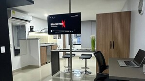 Une chambre d’hôtel moderne équipée d’un lit, d’un bureau avec un ordinateur portable, d’un téléviseur fixé au mur, d’un coin repas avec une table et des chaises, et d’une kitchenette à l’arrière-plan.