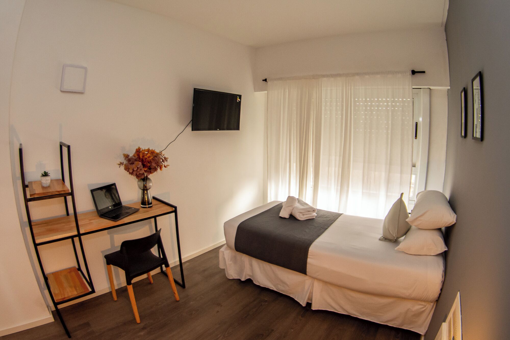 Een hotelkamer met een bed, een bureau met een laptop, een stoel, een vaas met bloemen en een aan de muur bevestigde televisie.