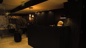 Zona de recepción con mostrador oscuro, una planta en maceta y pared de azulejos.