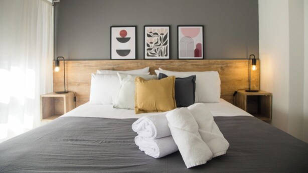 Una cama bien hecha con ropa de cama blanca y gris, una variedad de almohadas y un juego de toallas sobre la cama. Hay dos mesitas de noche con lámparas, y tres obras de arte enmarcadas en la pared sobre la cama.