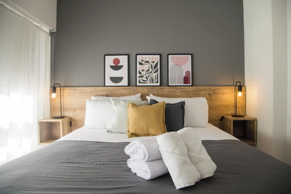 Un lit bien fait, avec du linge de lit blanc et gris, une variété d’oreillers et un ensemble de serviettes posées sur le lit. Il y a deux tables de chevet avec des lampes, et trois œuvres d’art encadrées accrochées au mur au-dessus du lit.