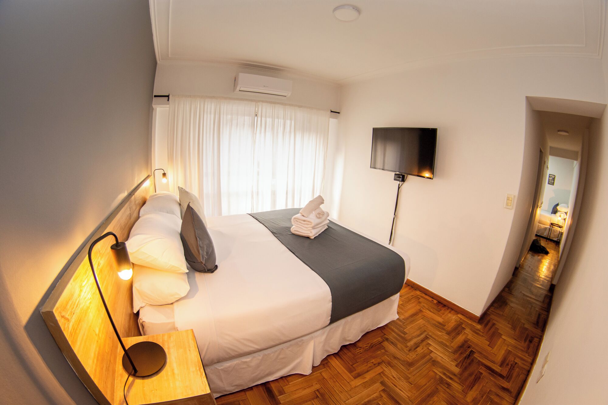 A hotel room with a bed, a flat-screen TV mounted on the wall, a bedside table with a lamp, and a wooden herringbone floor.