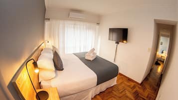 Habitación de hotel con cama, televisor de pantalla plana montado en la pared, mesita de noche con lámpara y suelo de madera en diseño espiga.