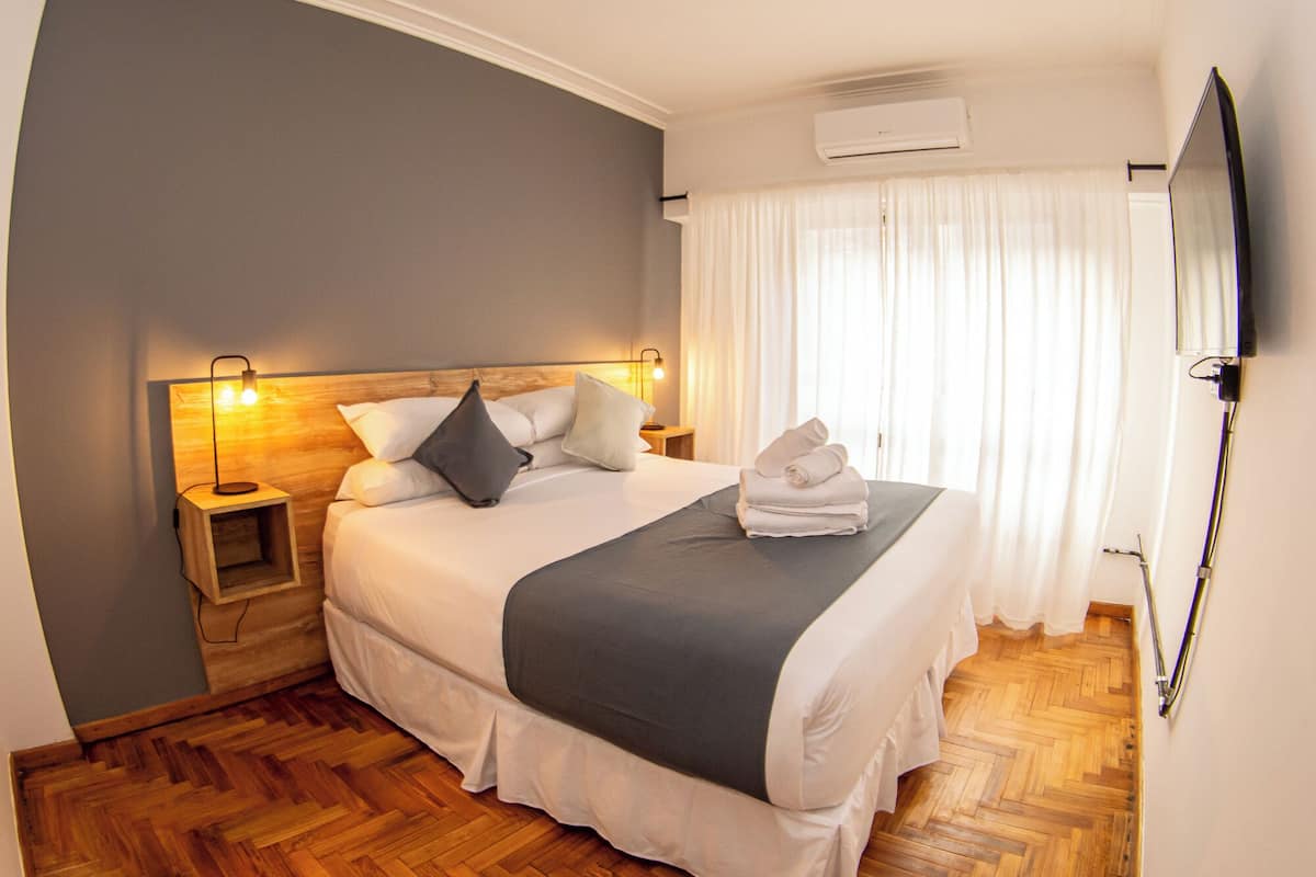 Un lit bien fait, recouvert d’une couverture grise, avec des oreillers blancs et une serviette pliée posée sur le côté. On y trouve une table de chevet en bois avec une lampe, un climatiseur fixé au mur et un téléviseur accroché au mur.