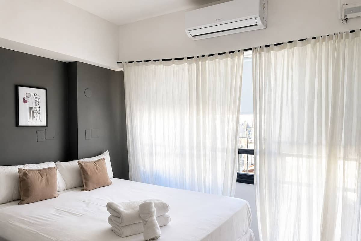 Une chambre d’hôtel moderne avec un grand lit, du linge de lit blanc, une œuvre d’art encadrée au mur et des rideaux en voile.