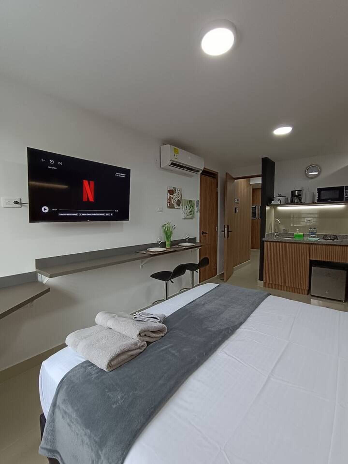 Moderni hotellihuone, jossa on sänky, seinälle asennettu televisio, pieni keittiönurkkaus ja pieni oleskelualue.