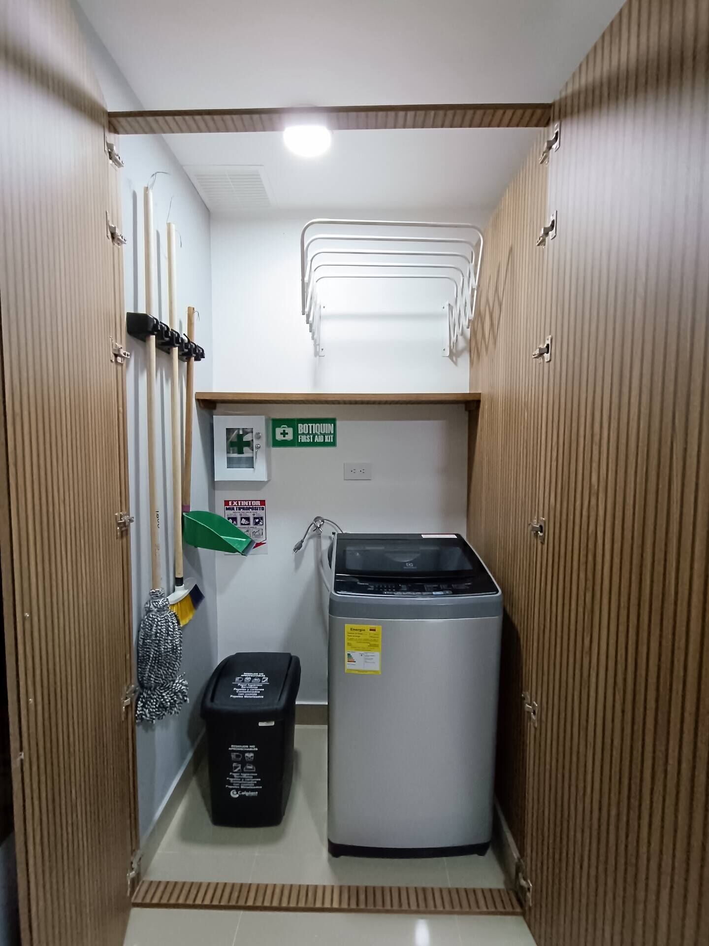 Ein kleiner, schmaler Raum mit einem Kühlschrank, einer Mülltonne und einem Schild für die Erste-Hilfe-Ausrüstung.
