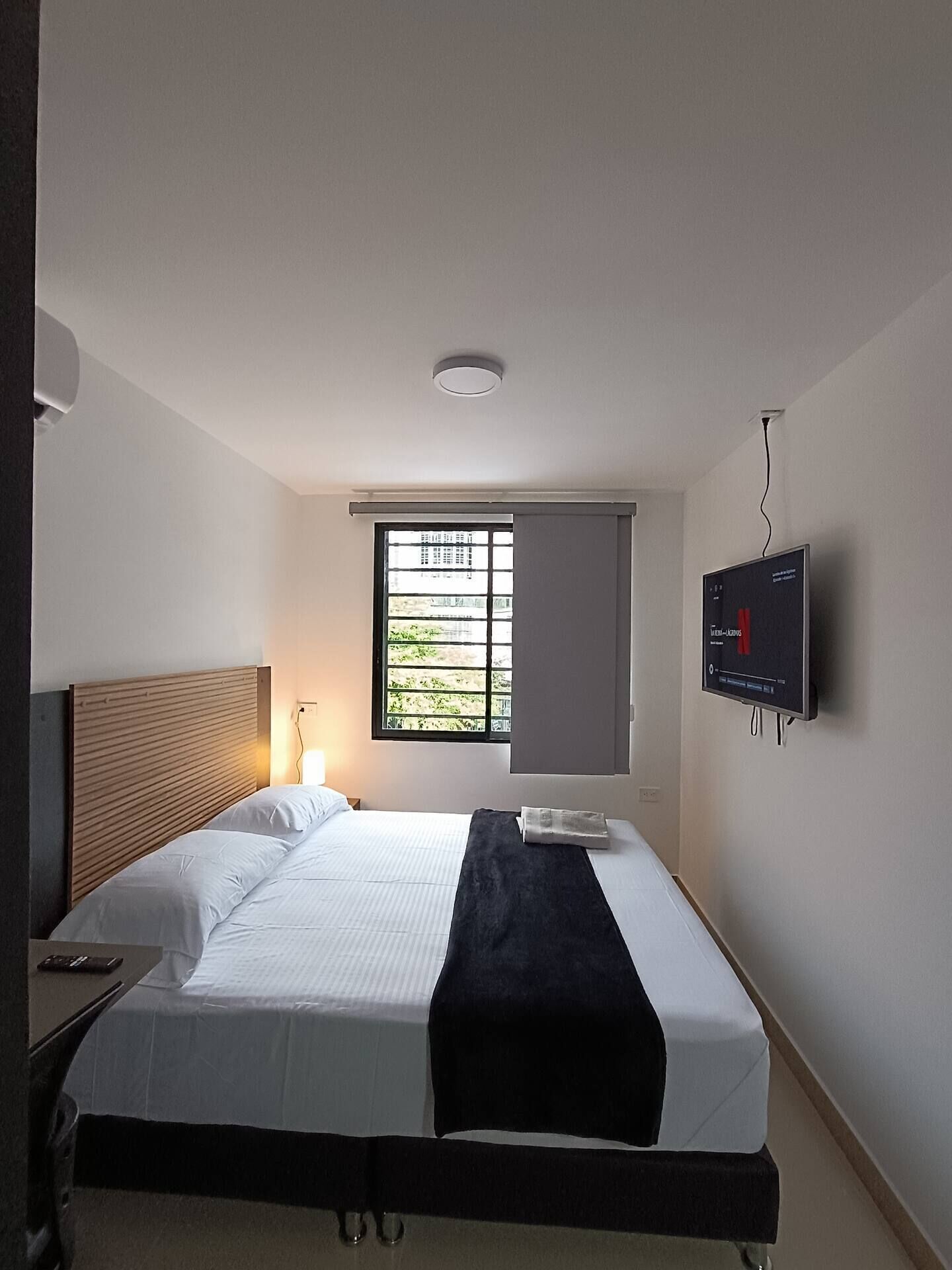 Une chambre d’hôtel avec un lit, un téléviseur fixé au mur, une fenêtre avec des stores et un luminaire au plafond.