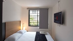 Een hotelkamer met een bed, een aan de muur bevestigde televisie, een raam met zonwering en een plafondlamp.