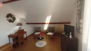 A small, cozy room with a wooden desk, two chairs, a small table with a vase of flowers, a television on a stand, and a wall-mounted shelf. - Guest rooms upstairs.  Breakfast included.  (Saillenard)
