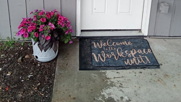 A white door with a frosted glass panel, a doormat with "Welcome to the Workspace unit" text, and a potted plant with pink flowers. - Workspace near Microsoft, Meta, Lumen Field and T-Mobile Park (Bellevue)