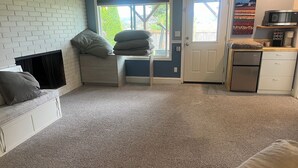 A compact living space with a white couch, a small bench with cushions, a kitchenette with a microwave and refrigerator, and a door leading to a garden. - Workspace near Microsoft, Meta, Lumen Field and T-Mobile Park (Bellevue)