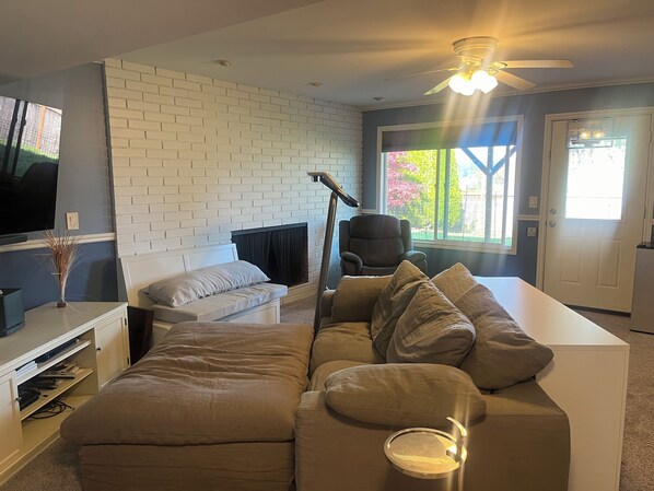 A living room with a white brick wall, a large window, a ceiling fan, and a flat-screen TV. - Workspace near Microsoft, Meta, Lumen Field and T-Mobile Park (Bellevue)