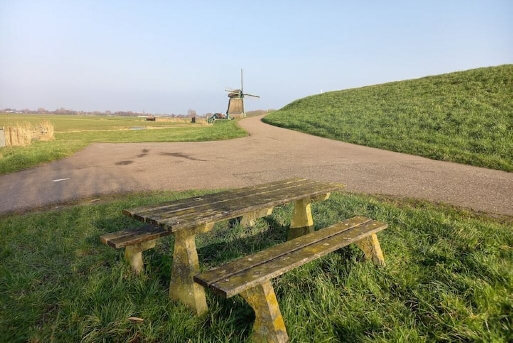 A windmill on a hill with a dirt path leading to it and a picnic table in the foreground.