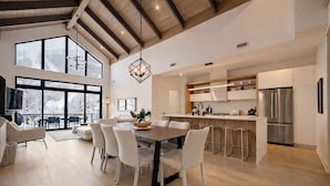 A spacious living area with a high ceiling, a dining table with chairs, a kitchen with modern appliances, and large windows offering a view of the outdoors.