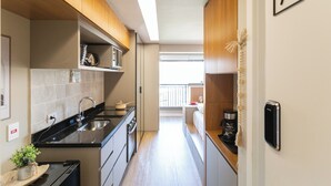 A narrow kitchen with wooden cabinets, a black countertop, and a window leading to a balcony. - Legacy Klabin (Sao Paulo)
