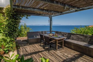 An outdoor patio with a wooden table set for dining, surrounded by cushioned seating, overlooking a scenic view of the sea and distant mountains.