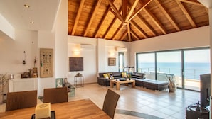 A spacious living area with a high wooden ceiling, a dining table with chairs, a sofa set, and a large window offering a view of the sea. - Sea Whisper Villa Peloponnese (North Kynouria)
