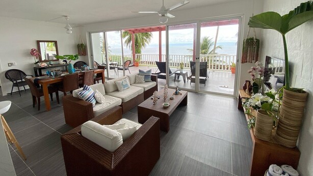 A spacious living area with a sofa set, dining table, and chairs. A balcony with a view of the ocean is visible through large sliding doors.