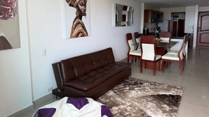 A modern living room with a brown leather sofa, a dining area with wooden chairs and a glass table, and a kitchen in the background. - Apartamento 8 personas 2 habitaciones  (Cartagena)