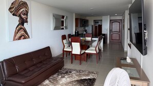 A modern living room with a brown leather sofa, a dining area with wooden chairs, a kitchen in the background, and a large flat-screen TV mounted on the wall. - Apartamento 8 personas 2 habitaciones  (Cartagena)
