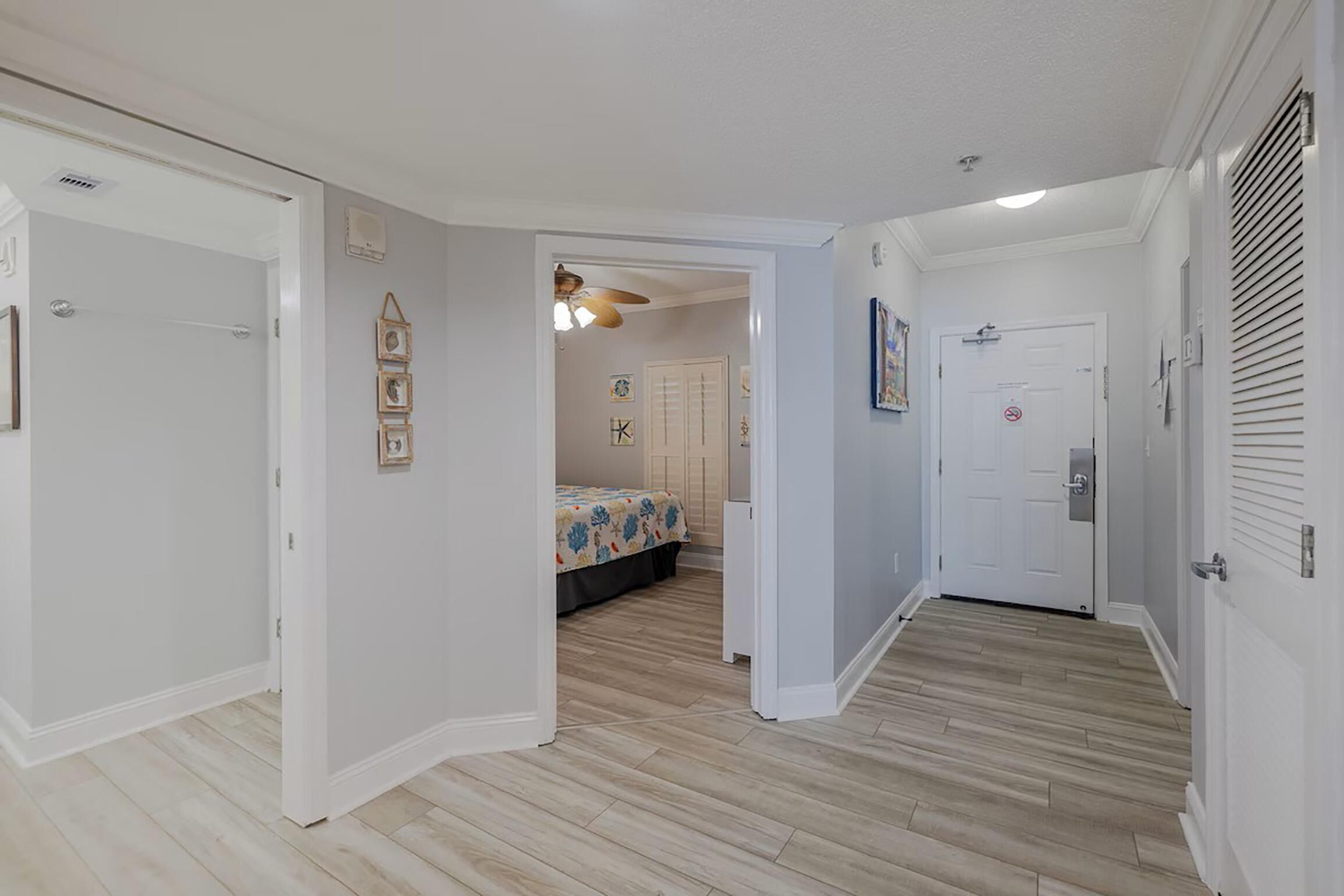 A hallway with wooden flooring, white walls, and a door leading to a bedroom with a bed and a ceiling fan.