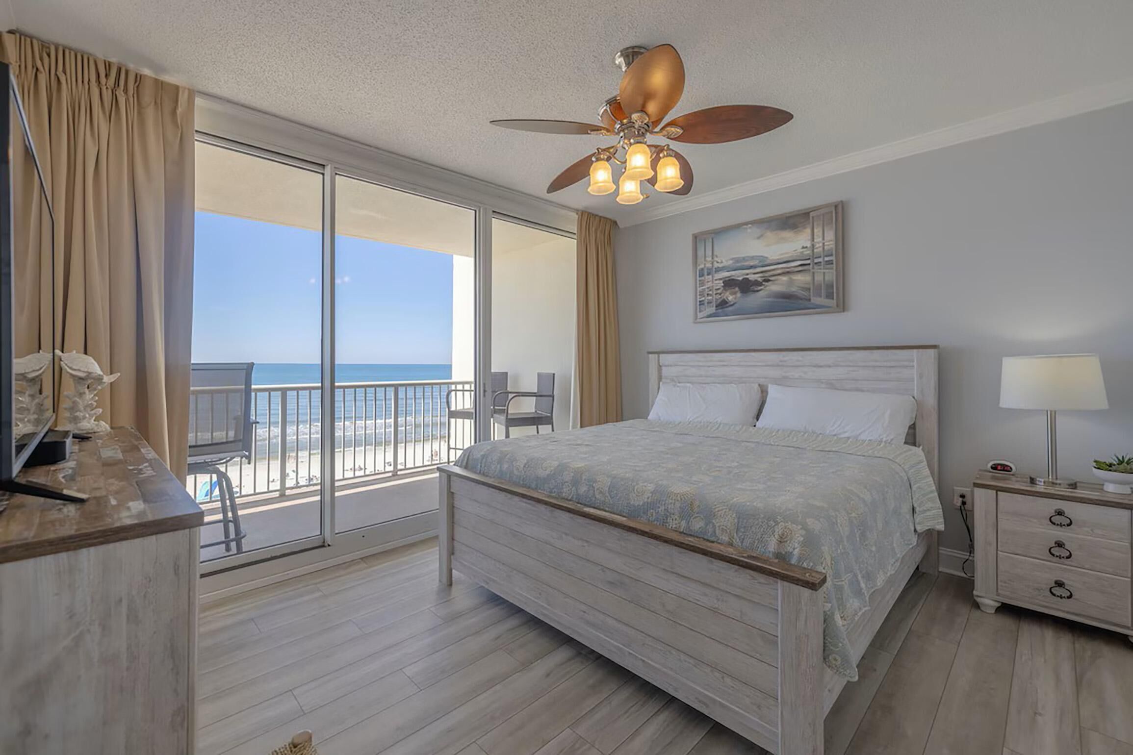 Un dormitorio con cama, mesita de noche, ventilador de techo, ventana con vista a la playa y una silla junto a la ventana.