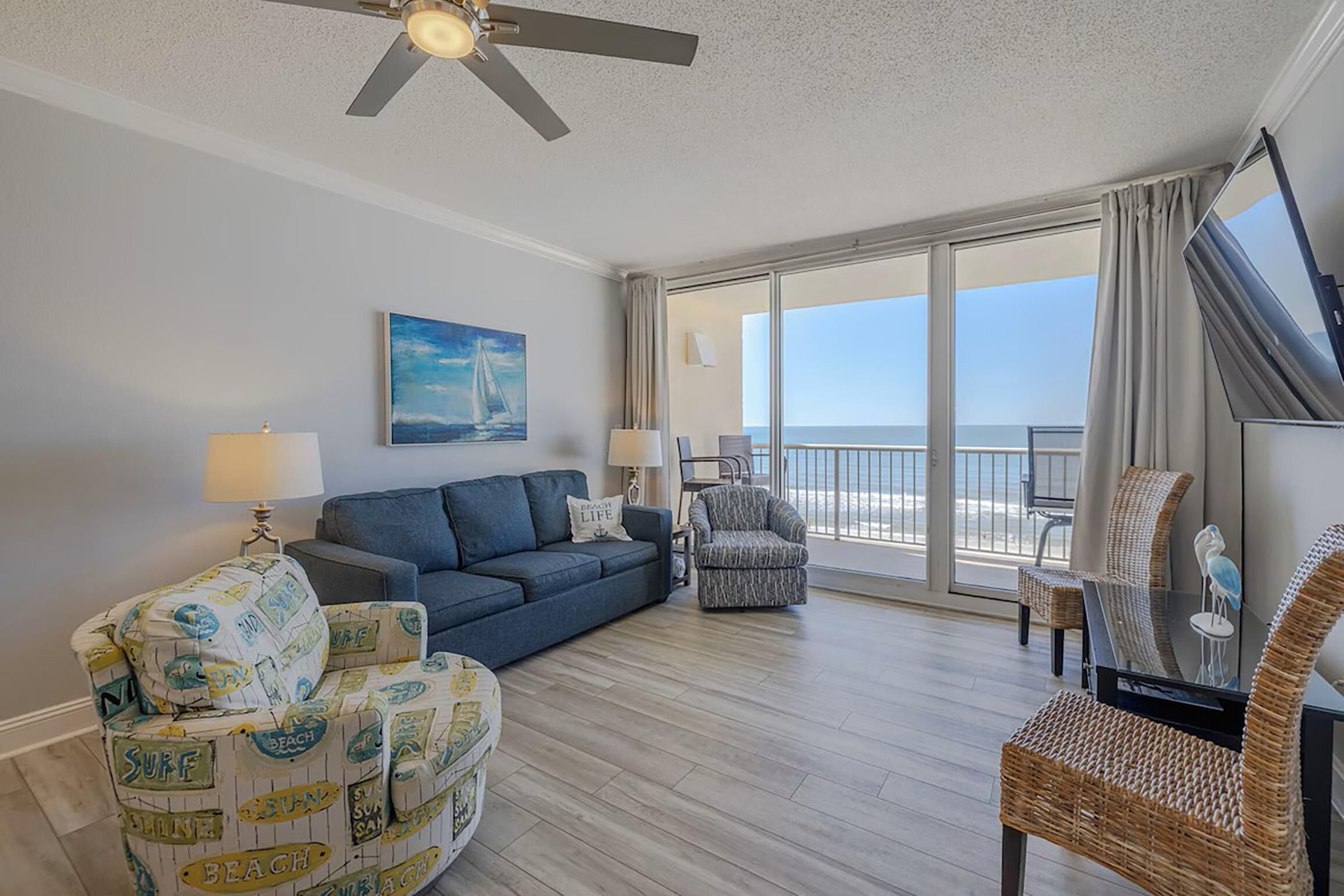 A spacious living room with a blue sofa, a patterned armchair, and a wicker chair. A ceiling fan is visible, and the room has a large window offering a view of the ocean.