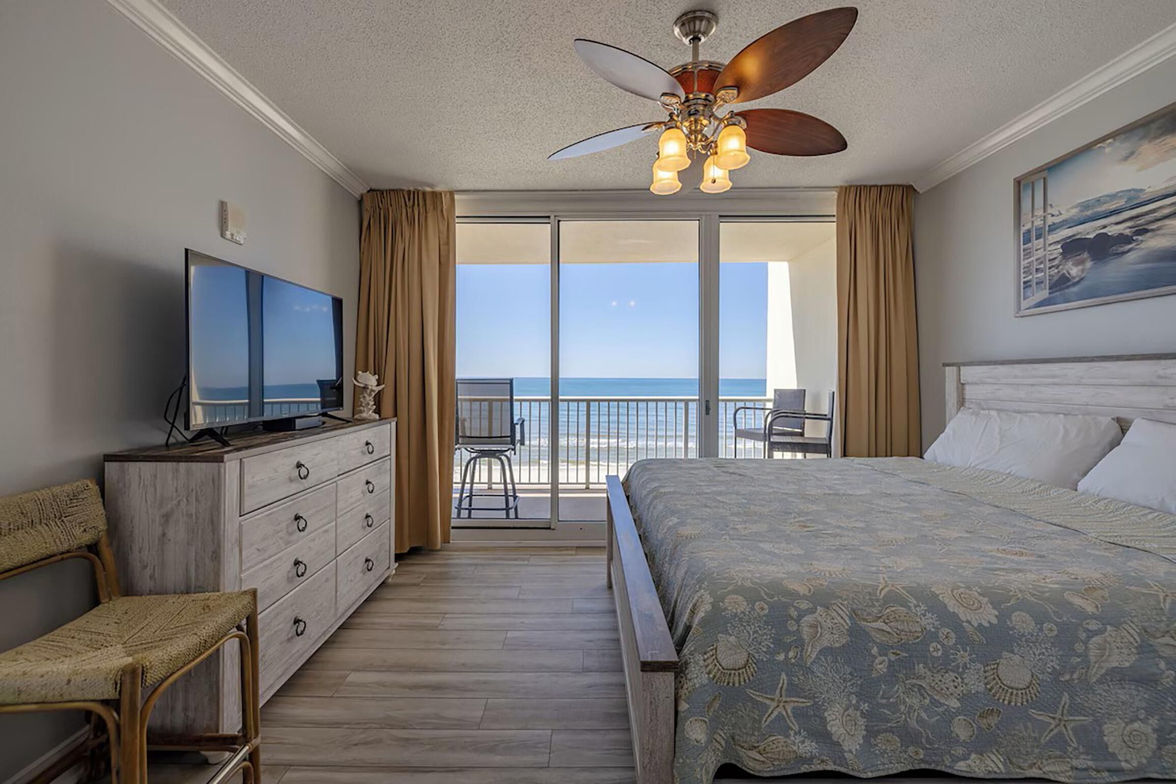 A hotel room with a bed, a television, a balcony with a view of the ocean, and a ceiling fan.