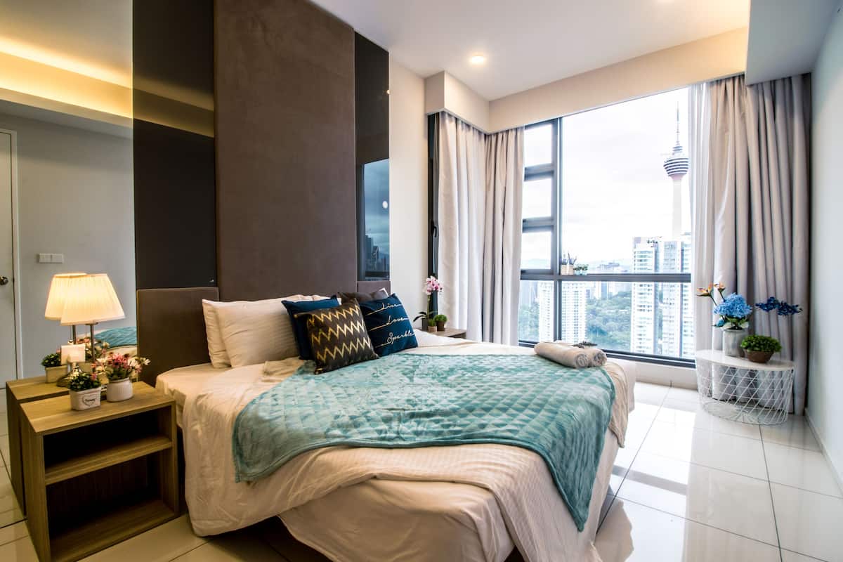 A modern hotel room with a large bed, a nightstand with a lamp, a view of the city, and a window with curtains.