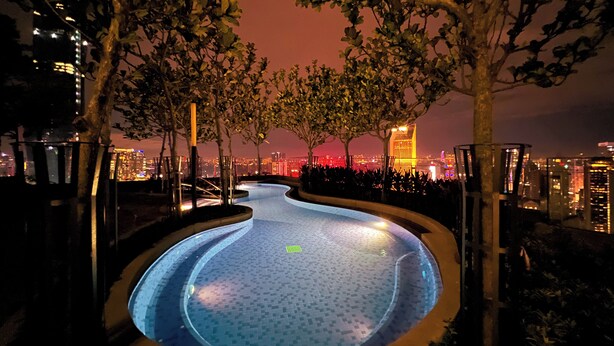 A nighttime swimming pool with a cityscape view, surrounded by trees and illuminated by artificial lighting.