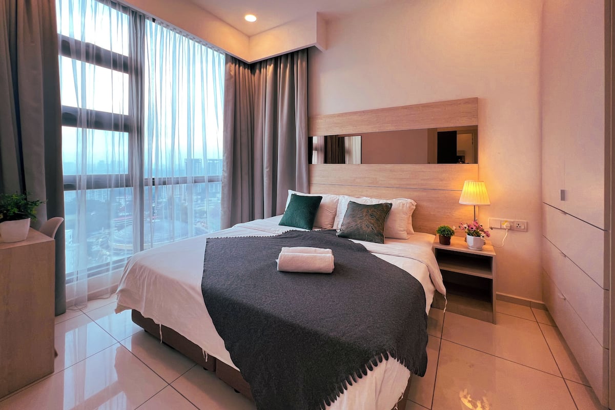 A modern hotel room with a large bed, a nightstand with a lamp, a potted plant, and a city view through large windows.