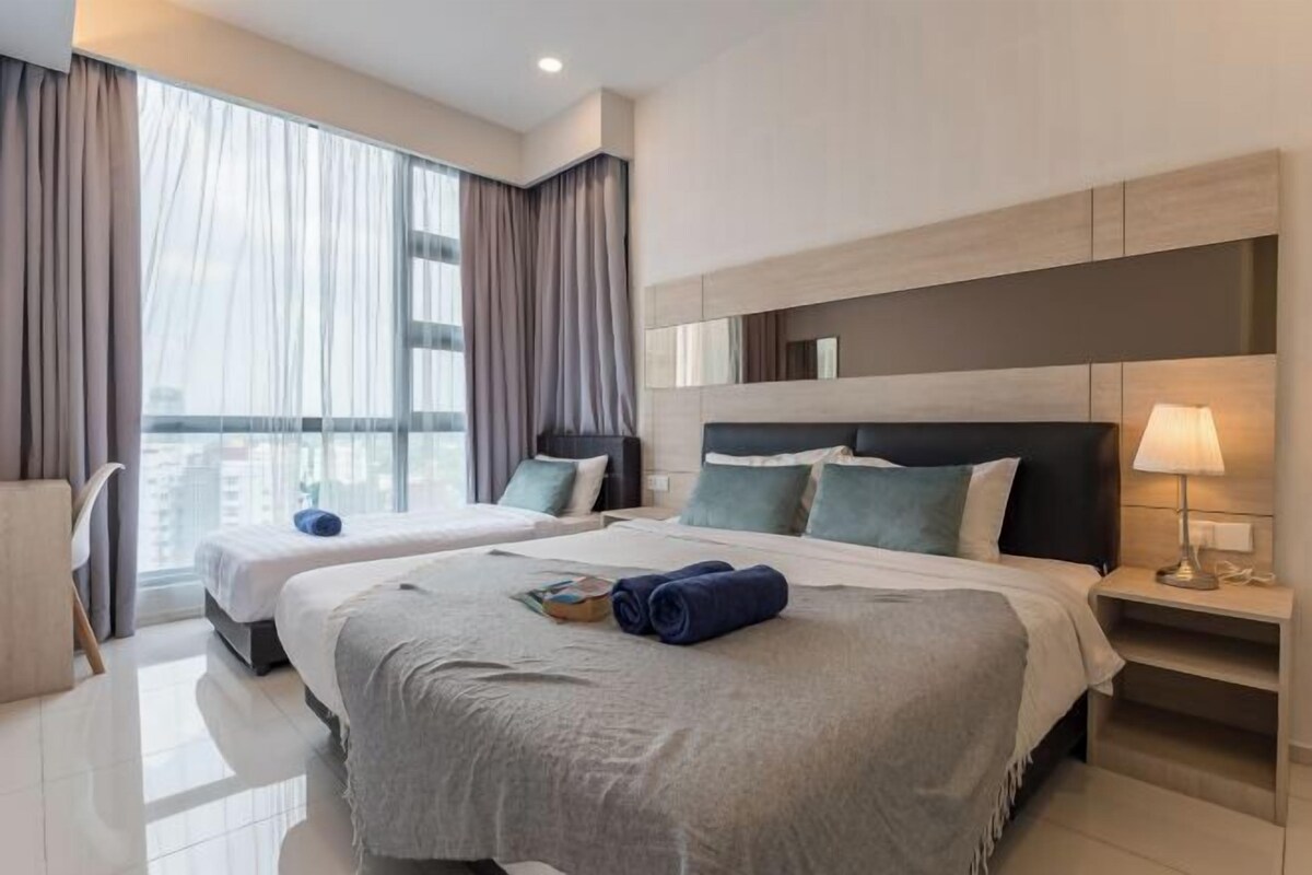 A hotel room with two beds, a large window with curtains, a bedside table with a lamp, and a view of the city.