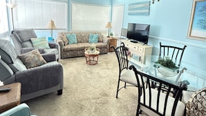 A living room with a ceiling fan, a glass dining table, and a sofa set. - Cute 2br Condo. Short walk to the beach. Close to shopping, restaurants & golf. (Myrtle Beach)