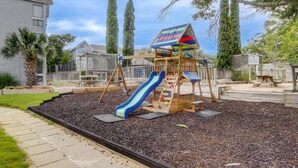 A playground with a slide and climbing area, surrounded by mulch and a fence. - Cute 2br Condo. Short walk to the beach. Close to shopping, restaurants & golf. (Myrtle Beach)